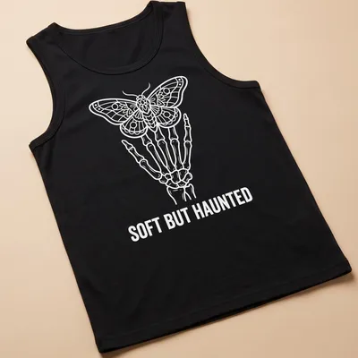 White line art of a skeleton hand holding a moth with the text Soft But Haunted — printed on a tank top