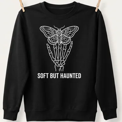 White line art of a skeleton hand holding a moth with the text Soft But Haunted — printed on a unisex sweatshirt