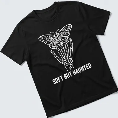 White line art of a skeleton hand holding a moth with the text Soft But Haunted