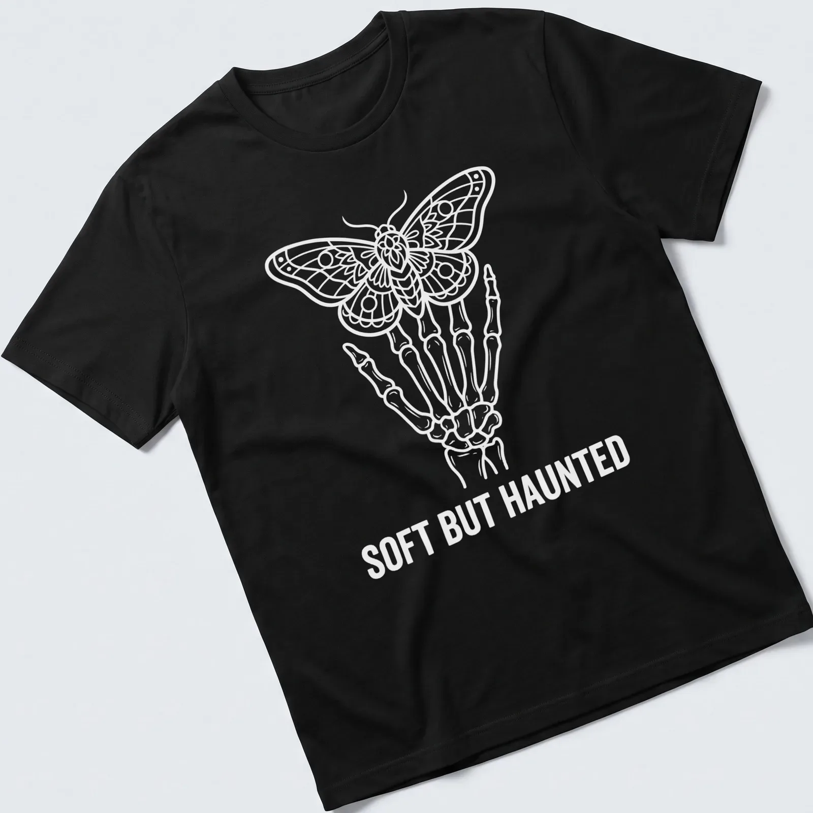 White line art of a skeleton hand holding a moth with the text Soft But Haunted — printed on a unisex t-shirt