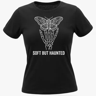 White line art of a skeleton hand holding a moth with the text Soft But Haunted — printed on a women's shirt