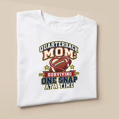 Bold, colorful text reads 'Quarterback Mom: Surviving One Snap at a Time' with football imagery — printed on a unisex t-shirt (angled view)