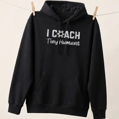 Bold playful text 'I COACH Tiny Humans' on a black and white soccer-themed — printed on a unisex hoodie