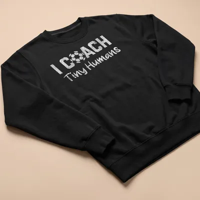 Bold playful text 'I COACH Tiny Humans' on a black and white soccer-themed — printed on a unisex sweatshirt
