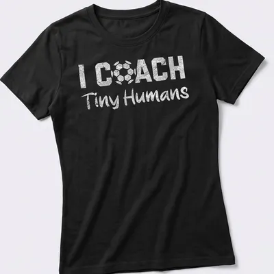 Bold playful text 'I COACH Tiny Humans' on a black and white soccer-themed — printed on a women's shirt